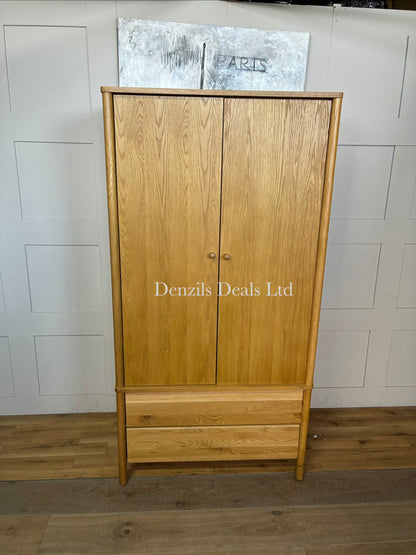 An elegant, mid-century modern freestanding double wardrobe crafted from warm, natural oak. The piece features two large doors with softer, rounded edges and clean minimalist vertical lines, opening to reveal a wooden hanging rail inside. Beneath the main cabinet section sit two wide, deep drawers stacked vertically for additional storage. The entire unit is accented by small, solid turned timber handles on both the doors and drawer fronts, giving it a smooth, unified, and contemporary look