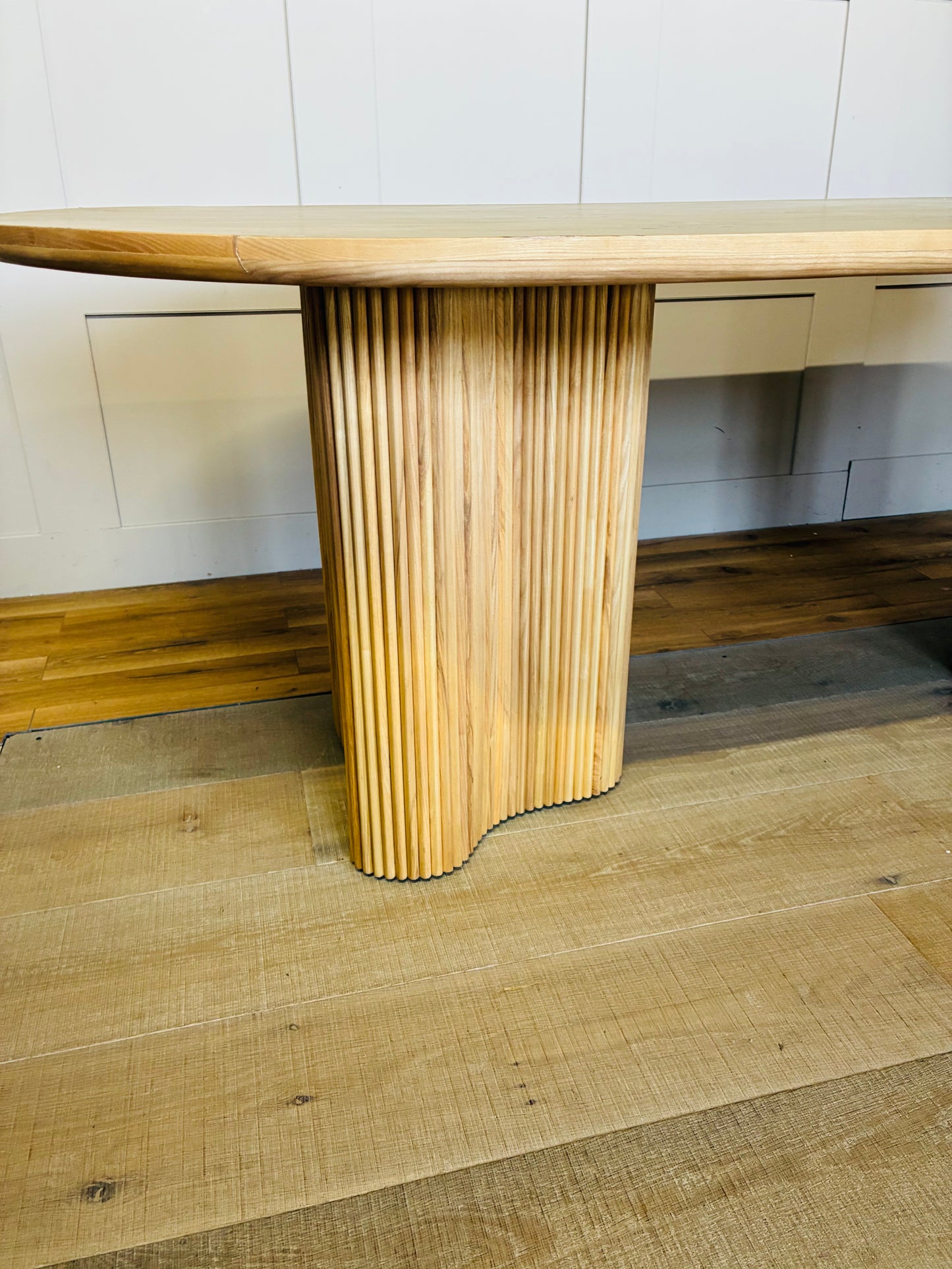 An Anthropologie Scout oval dining table made of light beige ash wood, featuring an oval tabletop with a decorative burl veneer pattern and thick, fluted pedestal legs. 