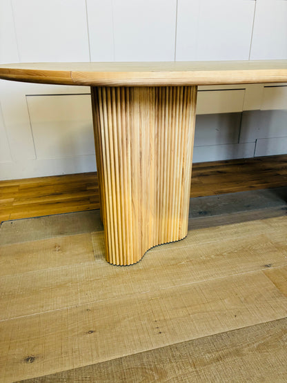 An Anthropologie Scout oval dining table made of light beige ash wood, featuring an oval tabletop with a decorative burl veneer pattern and thick, fluted pedestal legs. 