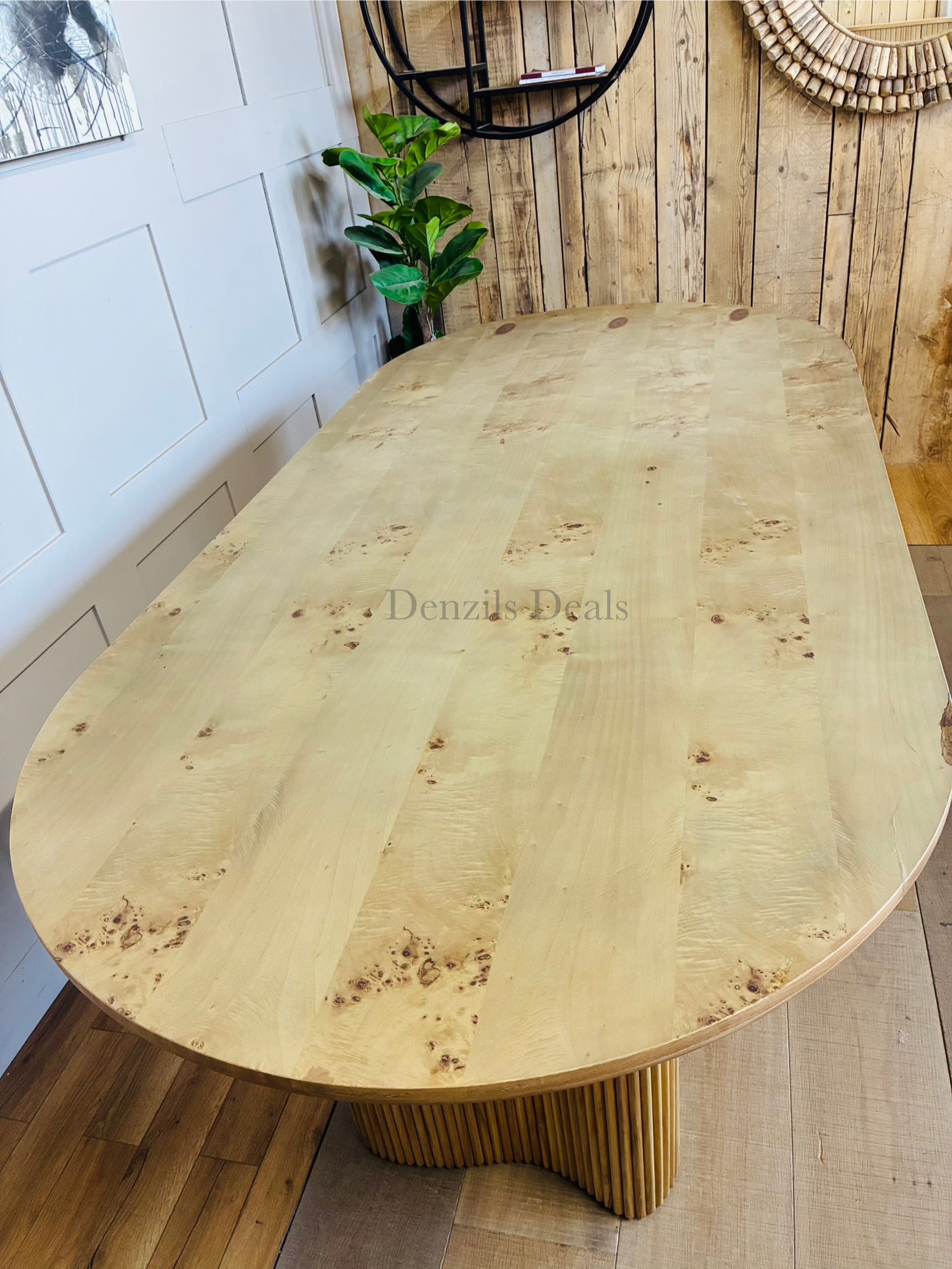 An Anthropologie Scout oval dining table made of light beige ash wood, featuring an oval tabletop with a decorative burl veneer pattern and thick, fluted pedestal legs. 
