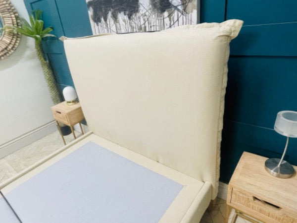 A contemporary double bed frame with a low-profile design. The headboard and frame are fully upholstered in a neutral stone-coloured fabric. It features vertical external seams on the corners and sits on four simple, angled wooden legs
