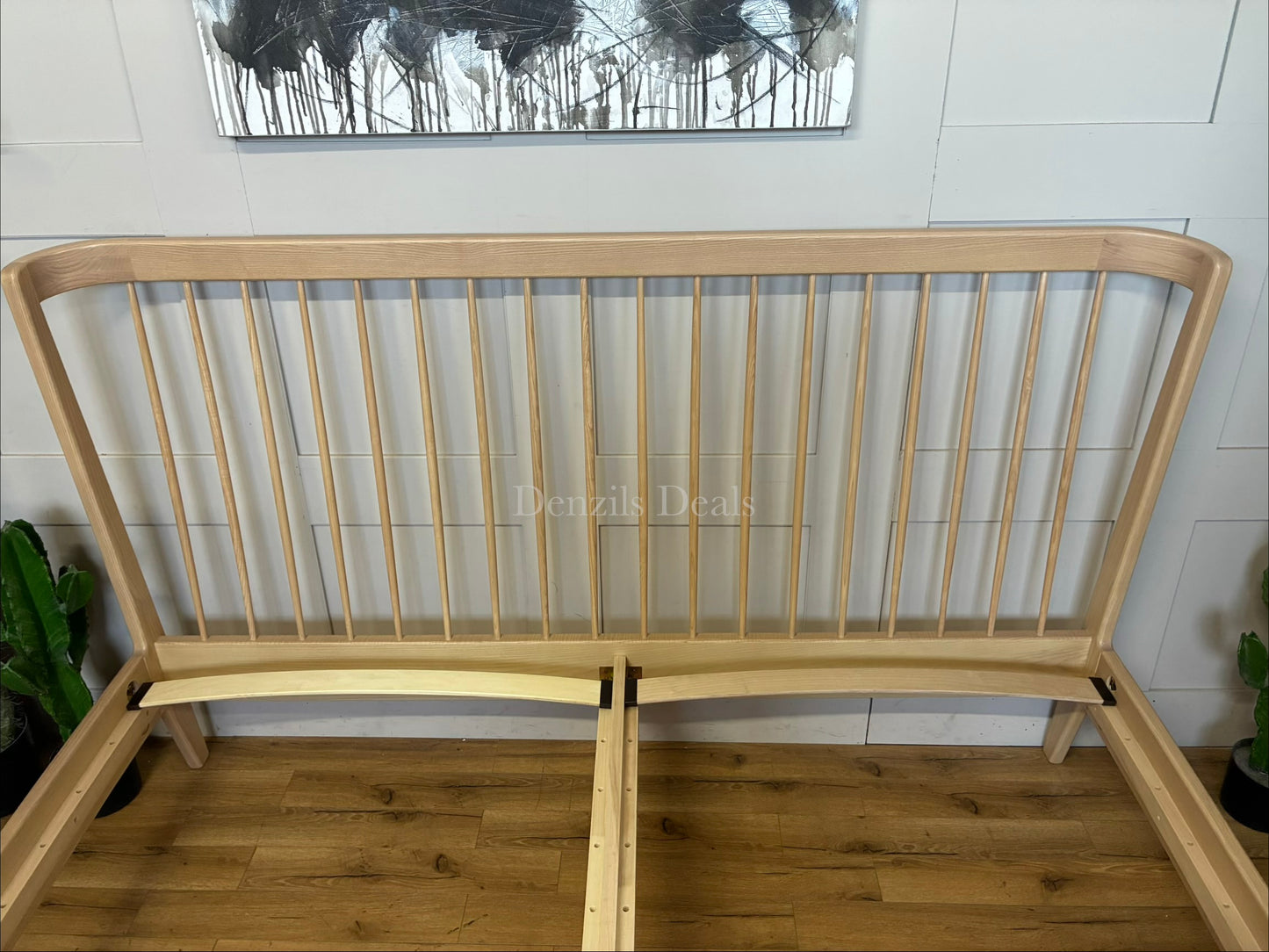 A super king-size bed frame made from natural-toned solid ash wood. The headboard has a distinctive curved silhouette with elegant turned spindles. The frame features softened profiles and fine veneers, supported by tapered wooden legs with 21.5cm of floor clearance