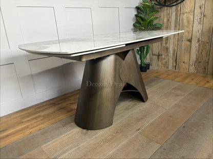 by a heavy-duty, sculptural metal pedestal base finished in a warm, matte bronze tone. The tabletop features a clean, seamless look with hidden internal tracking lines, illustrating its rotating extension mechanism that smoothly expands the surface from a compact six-seater to a larger eight-seater.