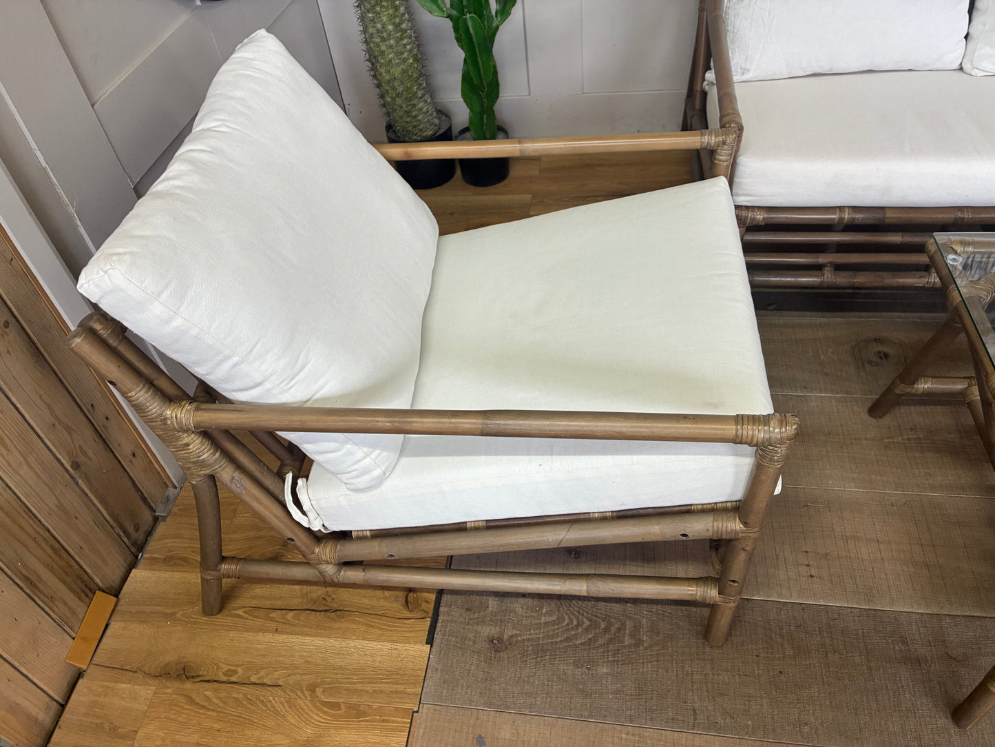 Four-piece bamboo lounge set in soil-brown finish, featuring a loveseat, two armchairs, and a matching bamboo coffee table with a glass top and bottom storage shelf. Thick, off-white cushions sit on open lattice bamboo frames