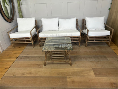 Four-piece bamboo lounge set in soil-brown finish, featuring a loveseat, two armchairs, and a matching bamboo coffee table with a glass top and bottom storage shelf. Thick, off-white cushions sit on open lattice bamboo frames