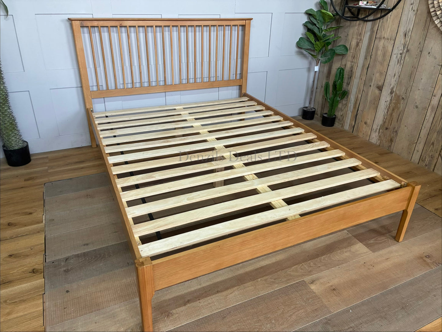 Front-angle view of the Julian Bowen Cotswold king-size bed frame in natural oak. The frame showcases a tall headboard with vertical wooden spindles, a low plain foot-end, robust corner posts, and an integrated wooden slatted base for mattress support.