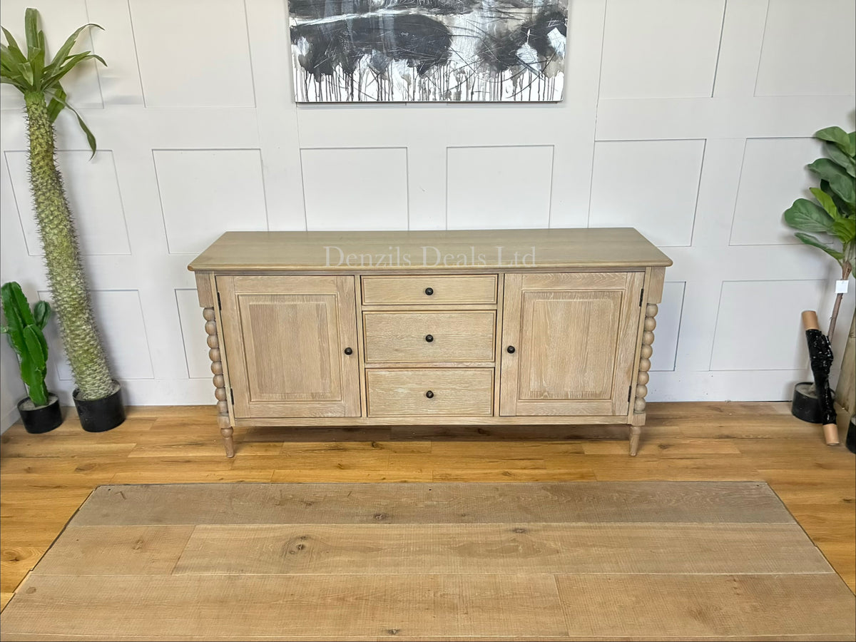 Gallery Direct Bessemer Oak Sideboard showcasing natural wood grains, smooth flat-front cabinetry, an overhanging top with curved edges, and distinctive artisanal hand-turned bobbin detailing trailing down each corner