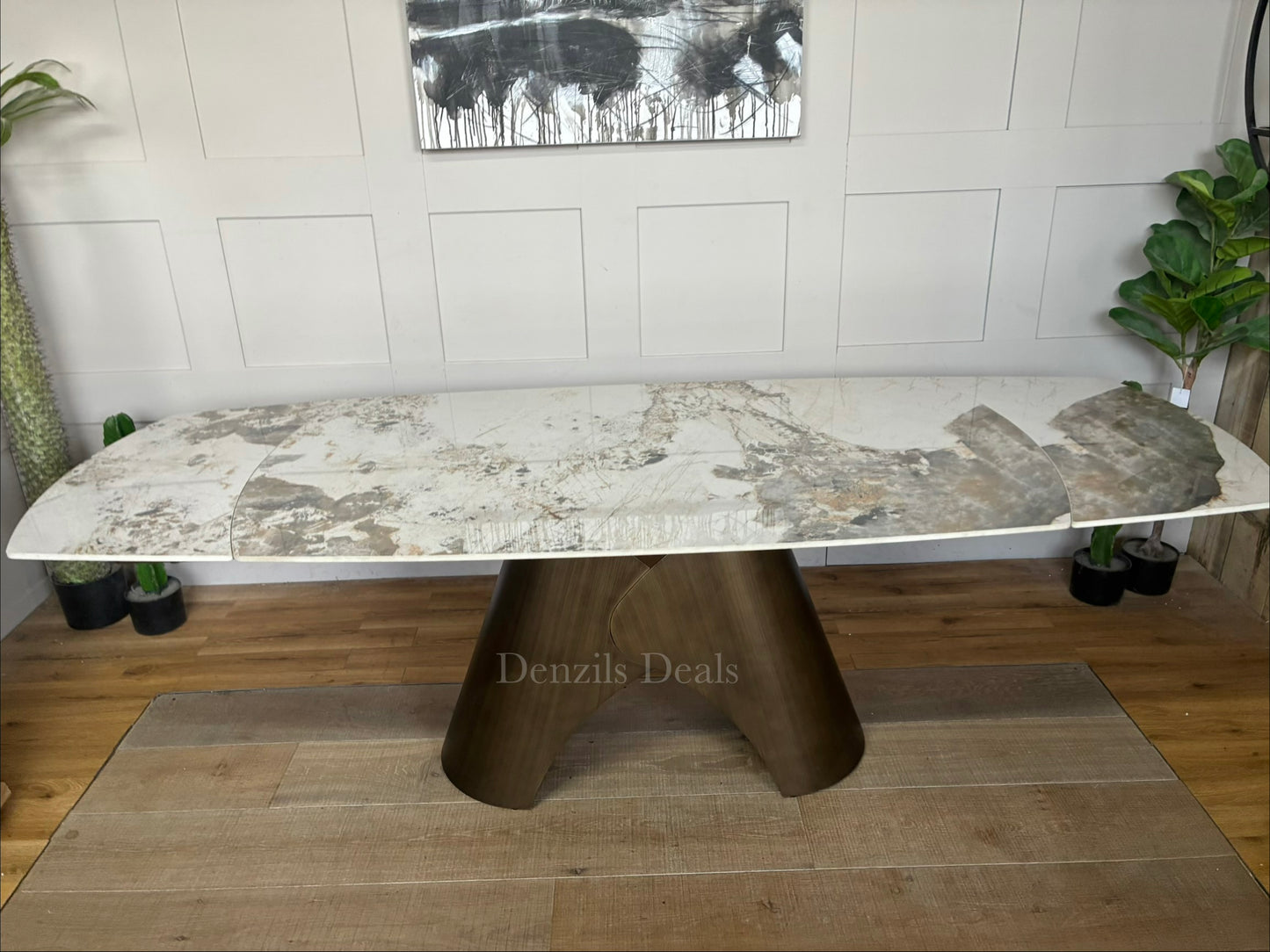 by a heavy-duty, sculptural metal pedestal base finished in a warm, matte bronze tone. The tabletop features a clean, seamless look with hidden internal tracking lines, illustrating its rotating extension mechanism that smoothly expands the surface from a compact six-seater to a larger eight-seater.