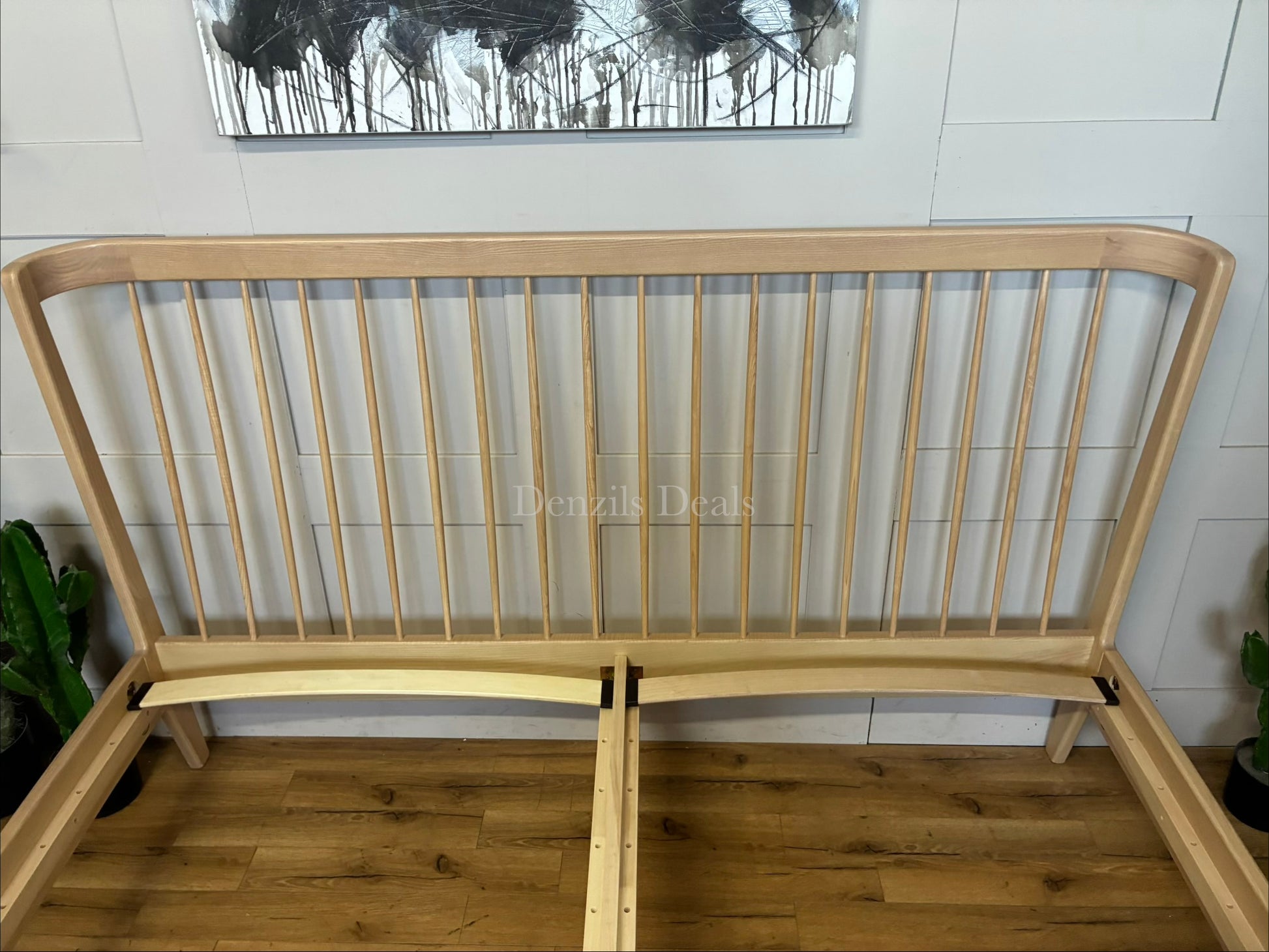 A super king-size bed frame made from natural-toned solid ash wood. The headboard has a distinctive curved silhouette with elegant turned spindles. The frame features softened profiles and fine veneers, supported by tapered wooden legs with 21.5cm of floor clearance