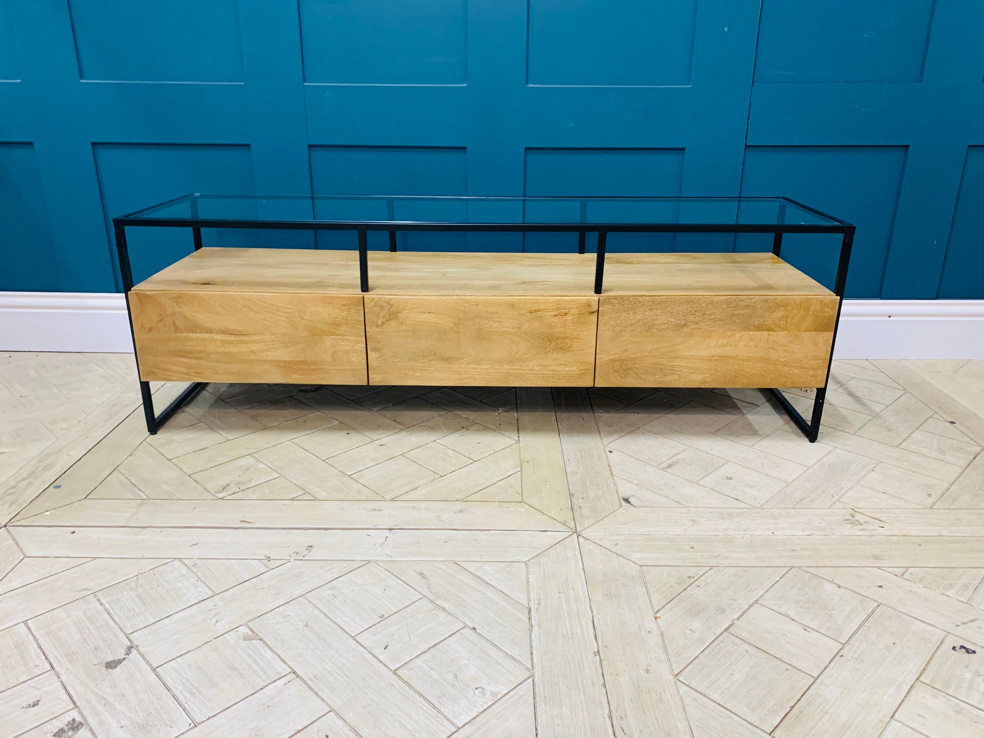 Industrial-style Made.com Kilby Wide TV stand. Features a warm, light mango wood grain base with two roomy storage drawers on clean, black metal legs. The top of the unit is a sleek smoked black glass, designed for placing a media console or television