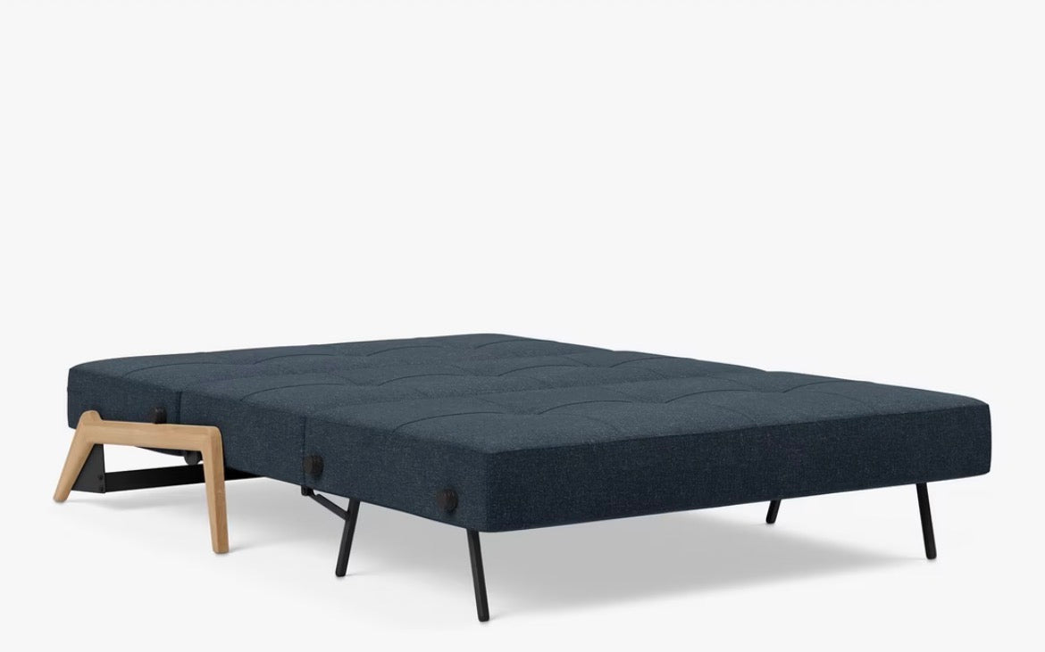The Cubed 140 sofa bed converted into a comfortable, modern double bed featuring a flat, supportive mattress in Nist Blue 515 fabric and exposed, light wooden or metallic supporting legs."