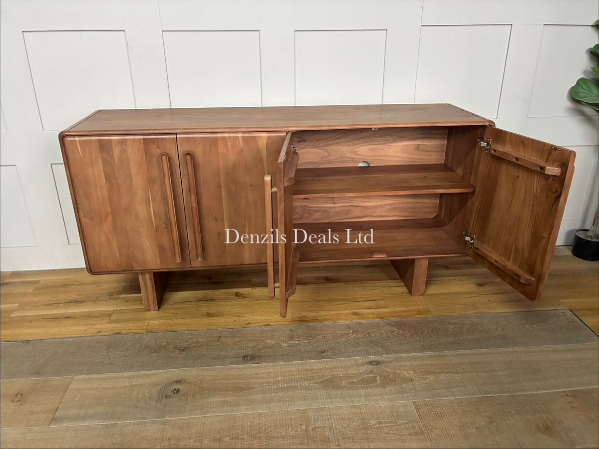 John Lewis Akola Sideboard in a light, natural wood tone. The sideboard is crafted from panelled acacia wood, creating vertical line patterns across its four minimalist doors. The outer edges of the frame are softly rounded, softening its blocky silhouette. The unit sits directly on the ground without legs, serving as a clean, mid-century or modern living room storage solution