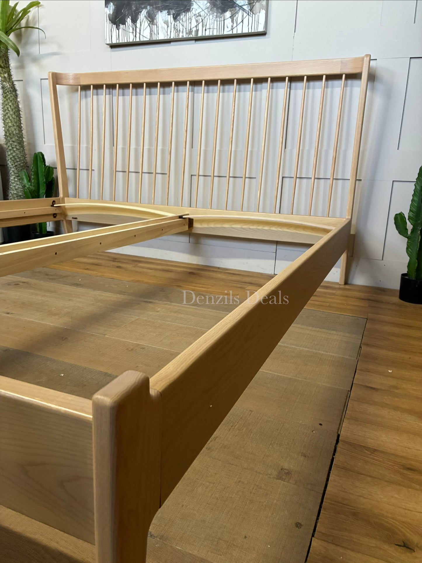 A super king-size bed frame made from natural-toned solid ash wood. The headboard has a distinctive curved silhouette with elegant turned spindles. The frame features softened profiles and fine veneers, supported by tapered wooden legs with 21.5cm of floor clearance