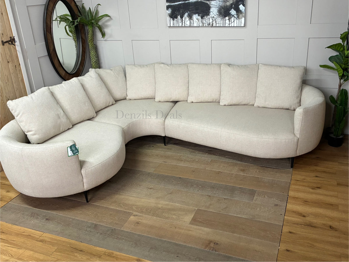 A contemporary 5-seater corner sofa by John Lewis. The sofa has a distinctive fluid design with soft, rounded organic edges. It is upholstered in a heavily textured, light beige 'twisted boucle' fabric in the shade 'Putty'. The unit curves into a left-hand facing configuration, supported by low-profile slim metal legs, and is topped with multiple matching square pillow cushions