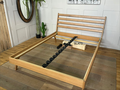A king-size minimalist platform bed frame made of light natural oak wood. It features a high, slatted headboard with clean horizontal lines, tapered angled legs, and a sturdy sprung-slat base.