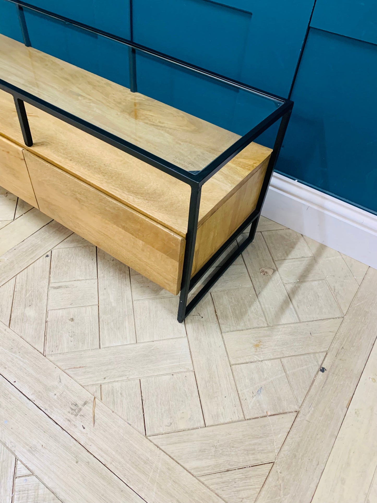 Industrial-style Made.com Kilby Wide TV stand. Features a warm, light mango wood grain base with two roomy storage drawers on clean, black metal legs. The top of the unit is a sleek smoked black glass, designed for placing a media console or television