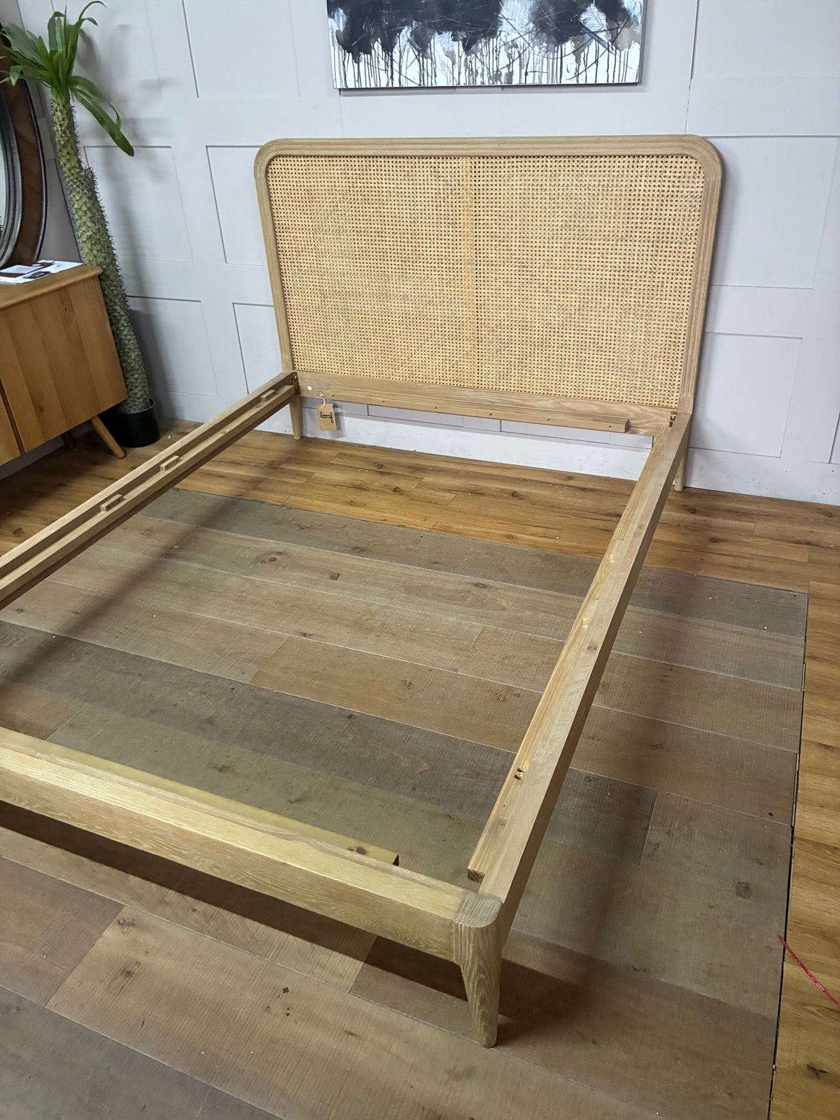 A light natural wood bed frame with a woven cane headboard, shown assembled on a wood plank floor in a room setting.