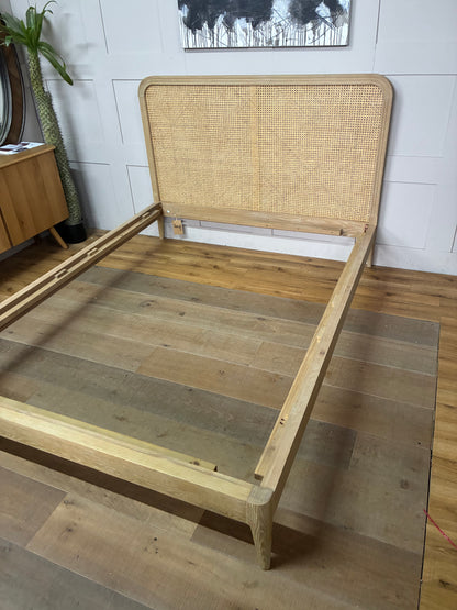 A light natural wood bed frame with a woven cane headboard, shown assembled on a wood plank floor in a room setting.