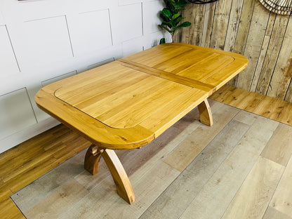 Large natural solid oak extendable dining table from Oak Furnitureland Hercules range featuring chunky cross-arched legs and chamfered beam.