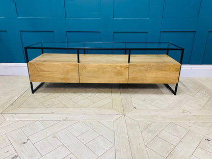 Industrial-style Made.com Kilby Wide TV stand. Features a warm, light mango wood grain base with two roomy storage drawers on clean, black metal legs. The top of the unit is a sleek smoked black glass, designed for placing a media console or television