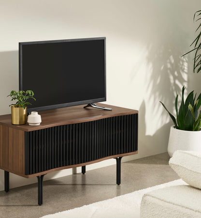 Made.com Zaragoza compact TV unit in organic walnut wood and charcoal black, featuring a contemporary slatted sliding front, subtle wavy edges, and slim metal legs
