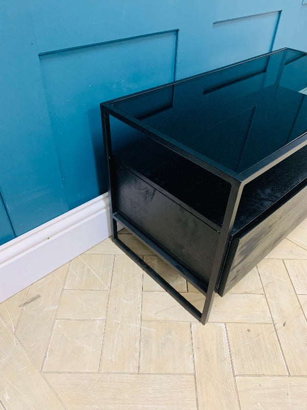 Modern Made.com Kilby TV stand featuring a solid mango wood base with a black stained finish. The unit includes a sleek single-drawer storage compartment and a smoked glass top, raised on slender, clean-lined metal legs