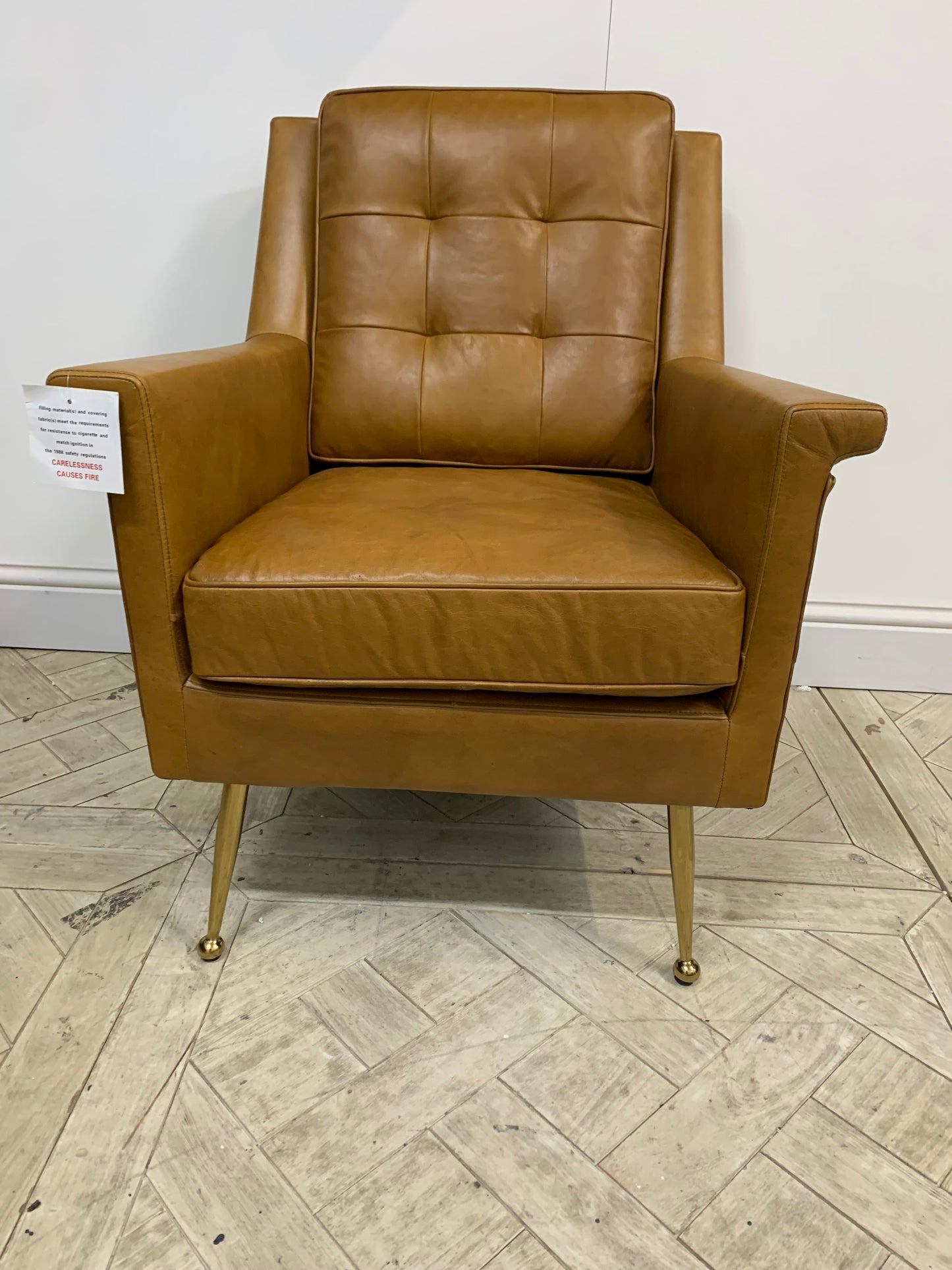Modern armchair in brown leather upholstery featuring buttoned back, folded details, and polished gold legs