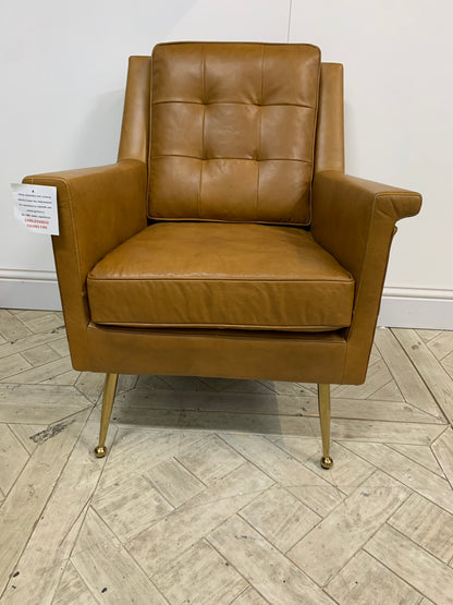 Modern armchair in brown leather upholstery featuring buttoned back, folded details, and polished gold legs