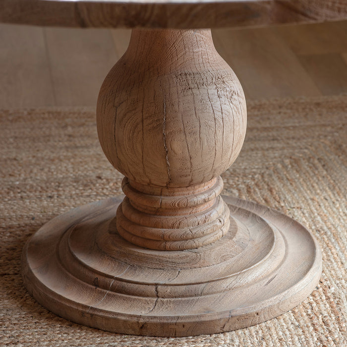 Nkuku Laya round light mango wood dining table featuring a thick circular top supported by a central, ornately carved baluster pedestal base with a tiered circular foot."