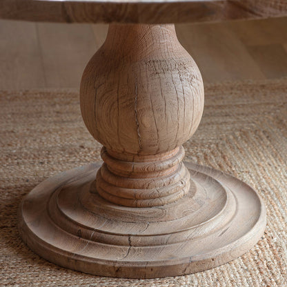 Nkuku Laya round light mango wood dining table featuring a thick circular top supported by a central, ornately carved baluster pedestal base with a tiered circular foot."
