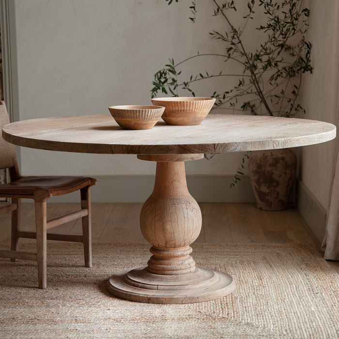 Nkuku Laya round light mango wood dining table featuring a thick circular top supported by a central, ornately carved baluster pedestal base with a tiered circular foot."