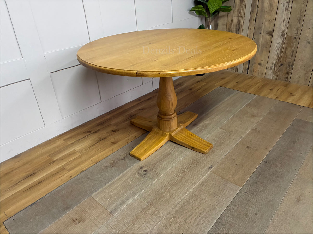Oak Furnitureland Henley range round dining table in natural oak. The image features a solid hardwood circular top with visible wood grains, supported by an ornate, chunky carved central pedestal column base resting on a stable cross-shaped foot