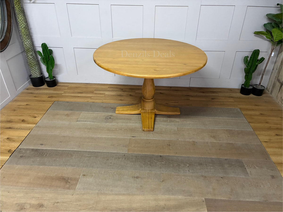Oak Furnitureland Henley range round dining table in natural oak. The image features a solid hardwood circular top with visible wood grains, supported by an ornate, chunky carved central pedestal column base resting on a stable cross-shaped foot