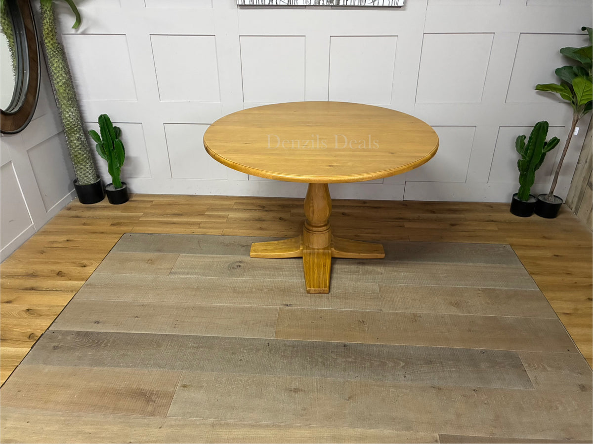 Oak Furnitureland Henley range round dining table in natural oak. The image features a solid hardwood circular top with visible wood grains, supported by an ornate, chunky carved central pedestal column base resting on a stable cross-shaped foot