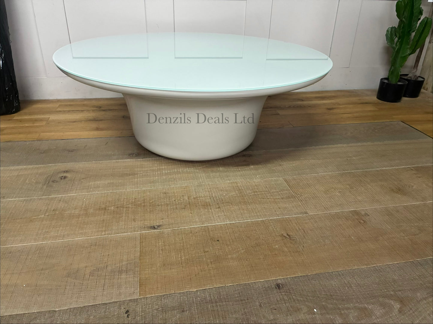 The modern Dulce 42” round coffee table with a glossy white top and a sculptural cylindrical pedestal base, set on a light brown wood plank floor against a white paneled wall.