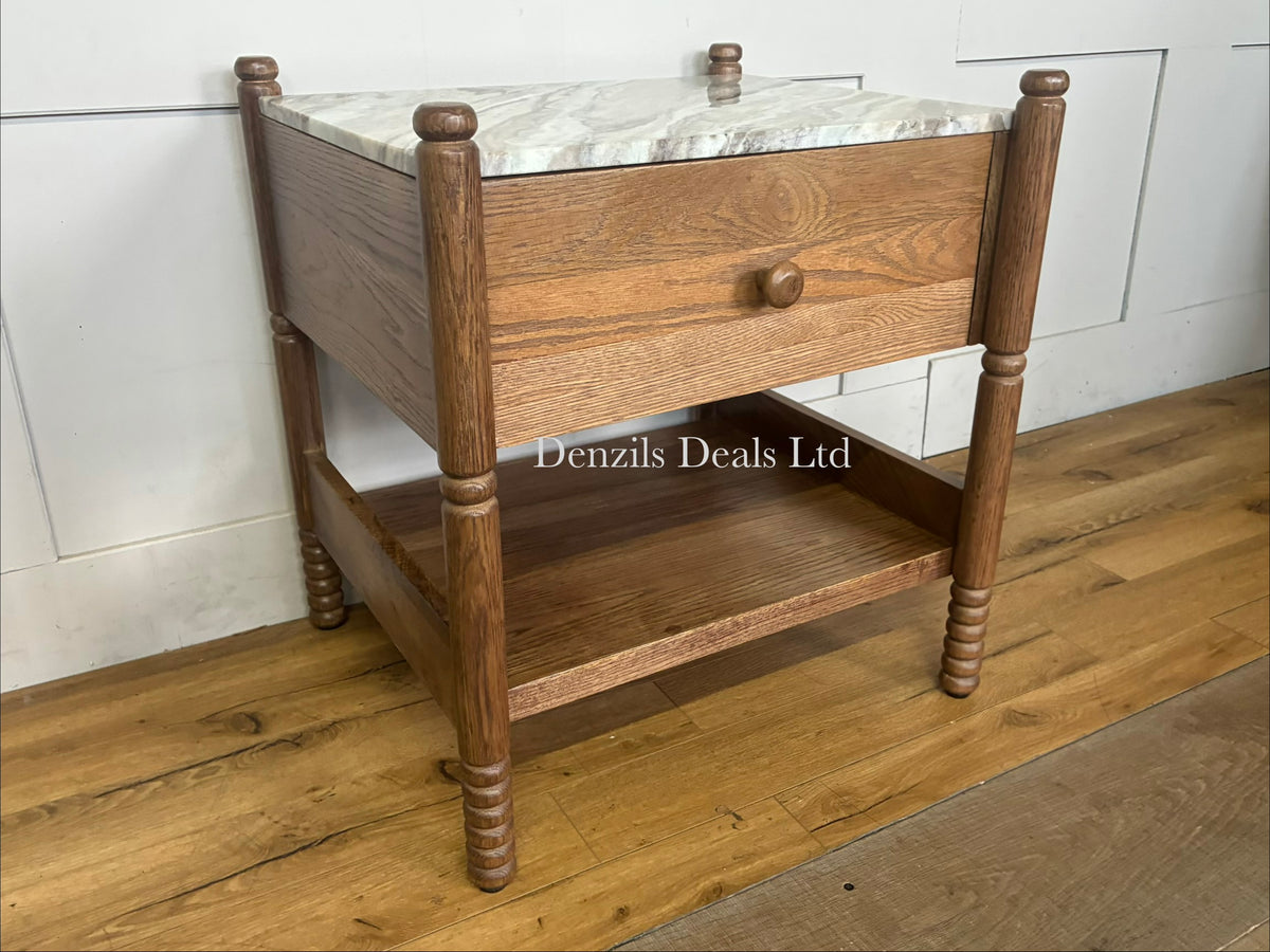 The Anthropologie Amelia Marble-Top Nightstand is a sophisticated bedside furniture piece that blends organic warmth with luxury materials. It features a premium Toronto marble top contrasted against an oak wood frame, complete with elegant spindled legs, a single storage drawer, and an open undermount shelf