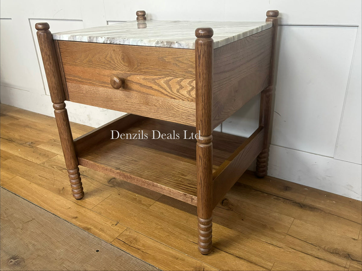 The Anthropologie Amelia Marble-Top Nightstand is a sophisticated bedside furniture piece that blends organic warmth with luxury materials. It features a premium Toronto marble top contrasted against an oak wood frame, complete with elegant spindled legs, a single storage drawer, and an open undermount shelf