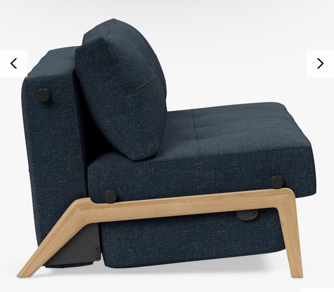 The Cubed 140 sofa bed converted into a comfortable, modern double bed featuring a flat, supportive mattress in Nist Blue 515 fabric and exposed, light wooden or metallic supporting legs."