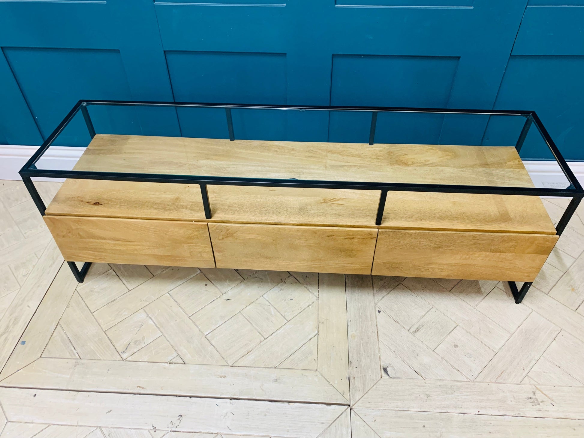 Industrial-style Made.com Kilby Wide TV stand. Features a warm, light mango wood grain base with two roomy storage drawers on clean, black metal legs. The top of the unit is a sleek smoked black glass, designed for placing a media console or television