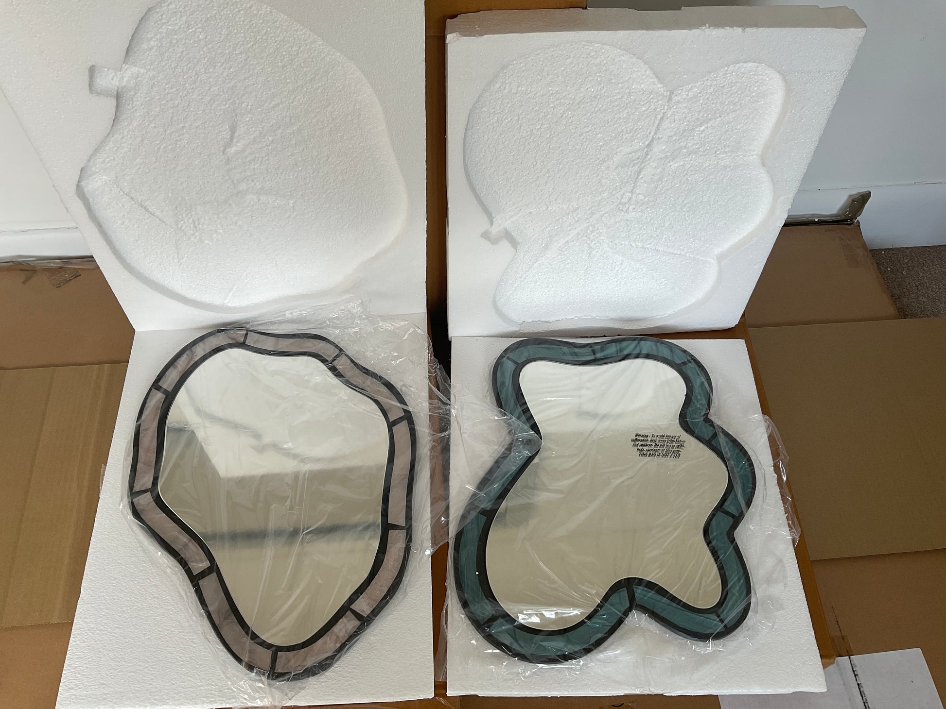 Two vertically oriented, rectangular wall mirrors featuring wavy, "squiggle" frames side-by-side. The mirror on the left has a light, pearlescent lilac purple frame, while the mirror on the right has a matching pearlescent soft blue frame, both displaying a subtle, glossy iridescence.