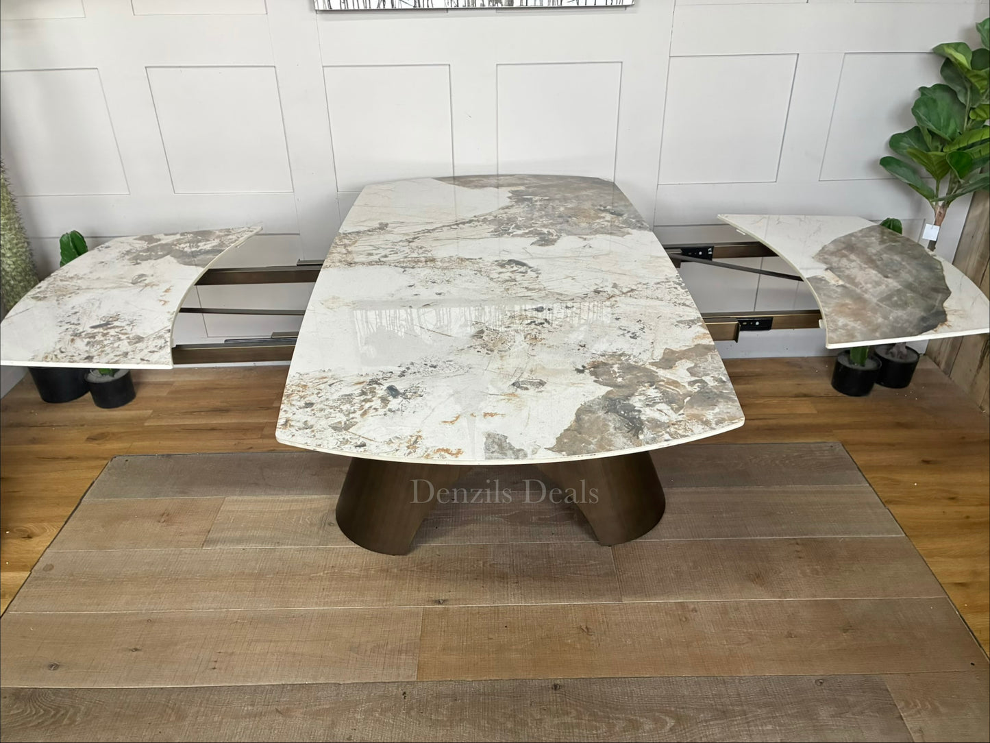 by a heavy-duty, sculptural metal pedestal base finished in a warm, matte bronze tone. The tabletop features a clean, seamless look with hidden internal tracking lines, illustrating its rotating extension mechanism that smoothly expands the surface from a compact six-seater to a larger eight-seater.