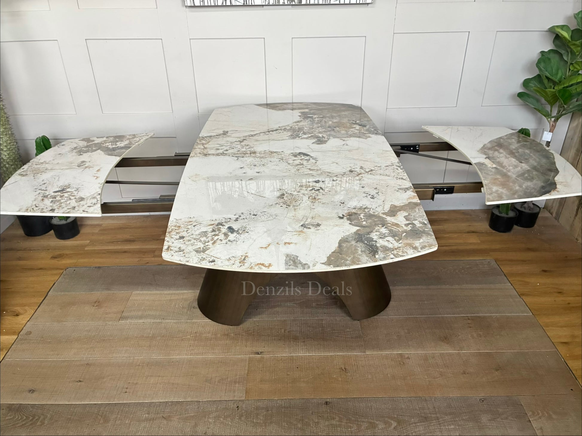 by a heavy-duty, sculptural metal pedestal base finished in a warm, matte bronze tone. The tabletop features a clean, seamless look with hidden internal tracking lines, illustrating its rotating extension mechanism that smoothly expands the surface from a compact six-seater to a larger eight-seater.