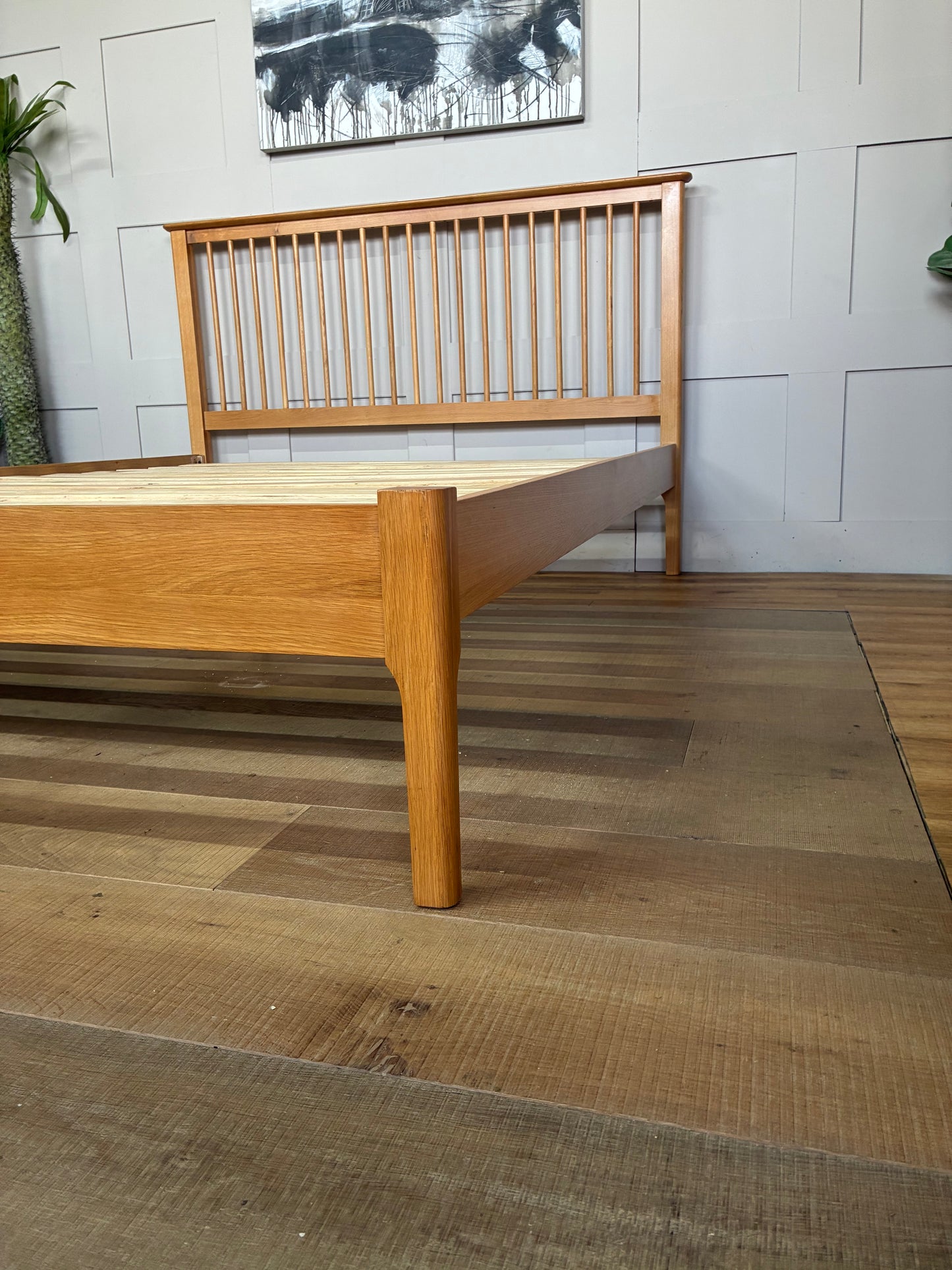 A king-size wooden bed frame made from light natural oak with a satin lacquer finish. The bed has a high headboard with vertical Shaker-style wooden slats, a low-profile matching footboard, and visible natural wood grain patterns