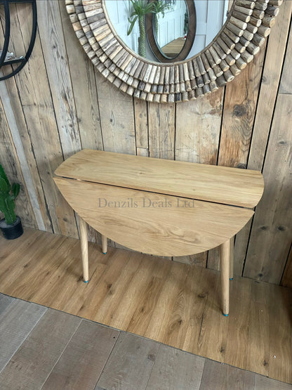 A round, space-saving wooden dining table with a smooth, light-toned natural oak veneer finish. The table is supported by a light wood frame featuring hinged gatelegs that swing out to hold up the folding drop leaves.