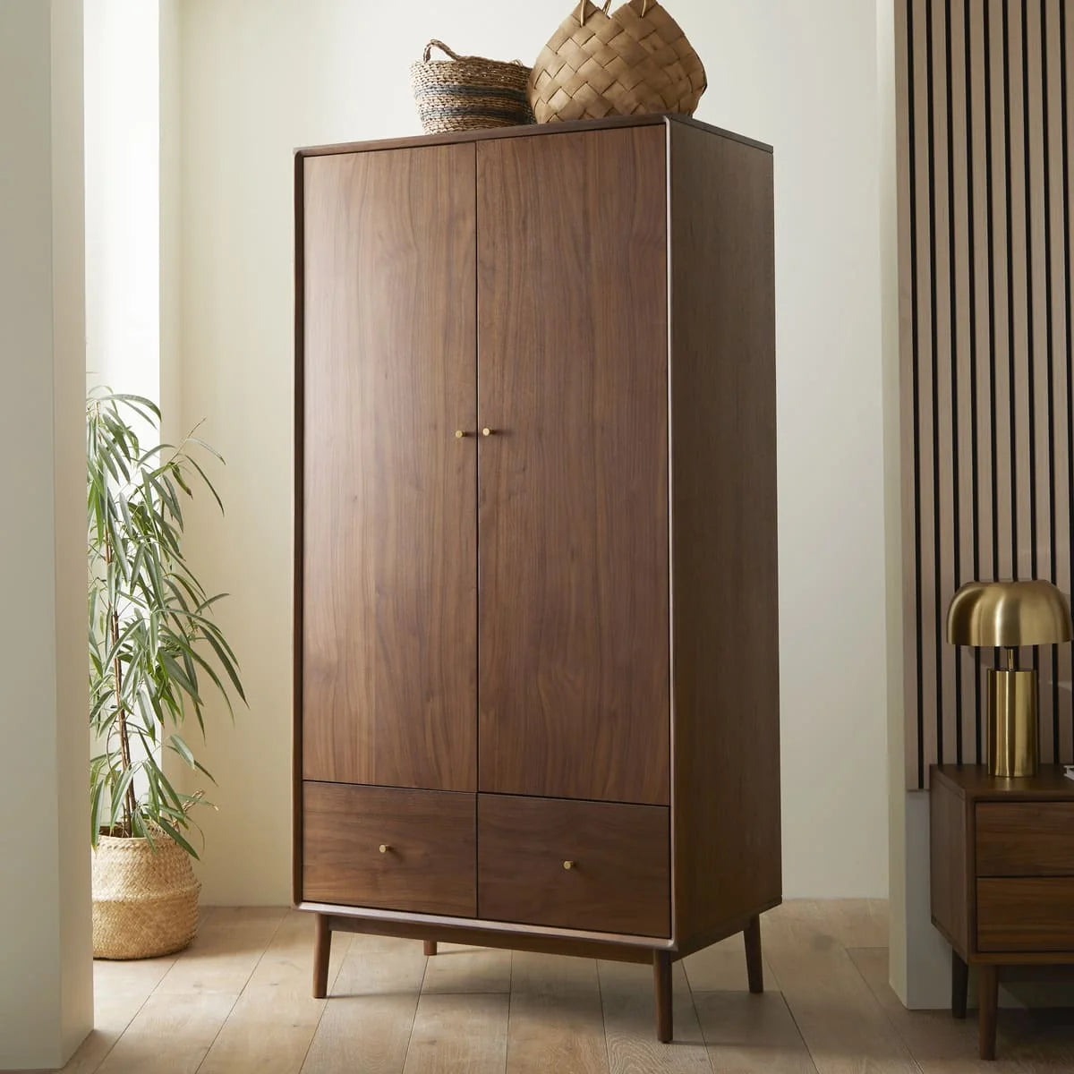 A mid-century modern two-door wooden wardrobe in a rich American walnut finish, featuring streamlined cabinet doors, polished brass knobs, and integrated base drawers